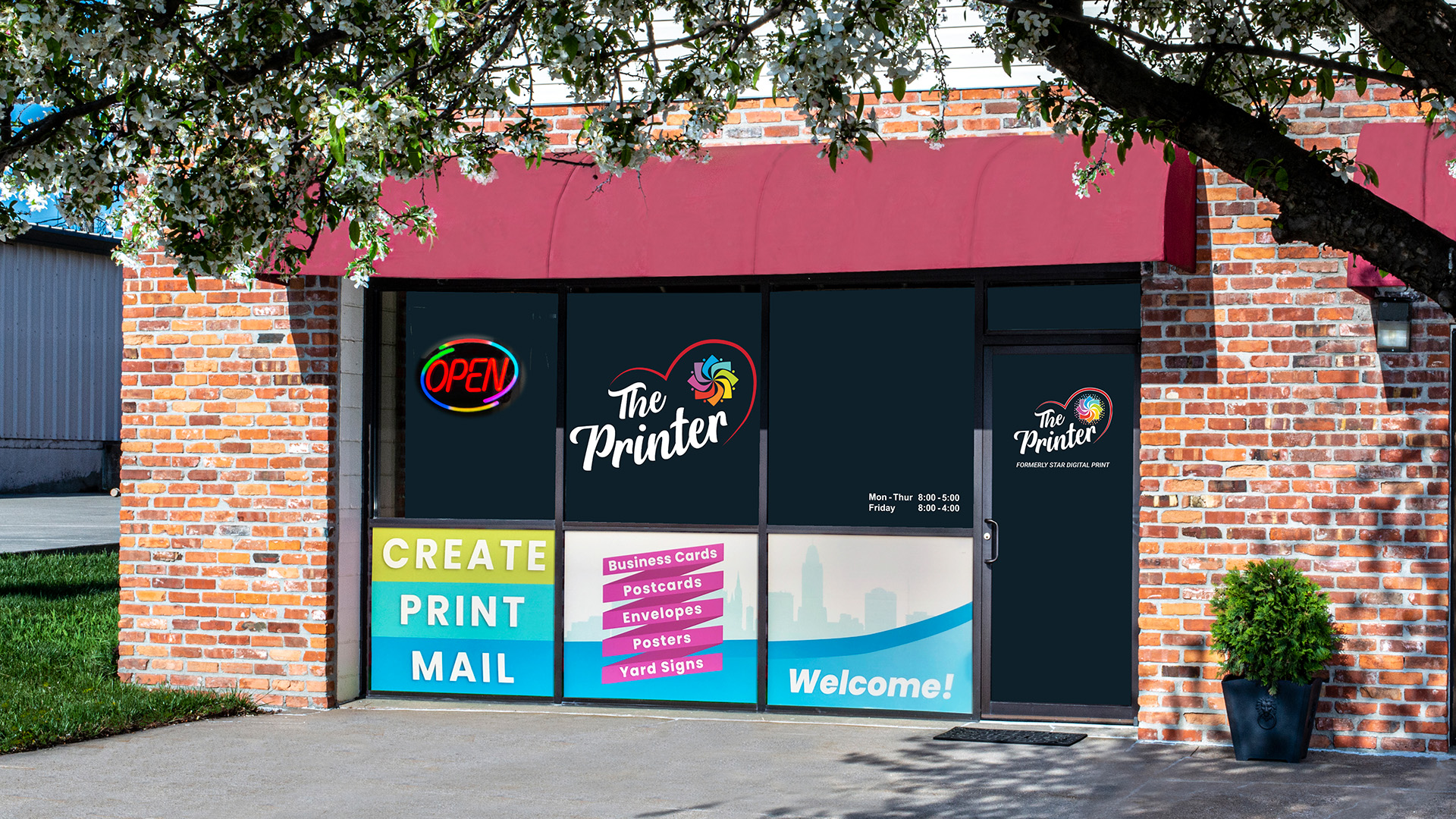Storefront of The Printer, featuring a red awning, brick exterior, and large windows with colourful signage advertising services like 'Create, Print, Mail' and business cards, postcards, and posters, with a neon 'Open' sign and a welcoming entrance.
